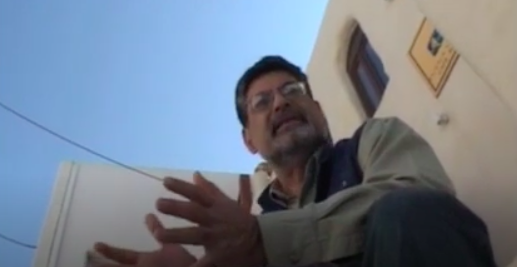 A man wearing glasses speaks in front of a building, the camera looks up from below