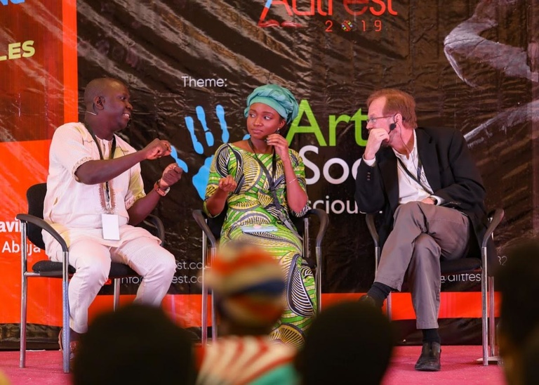 Three people engage in a panel discussion at ArtFest 2019. The event backdrop and audience members are visible in the foreground.