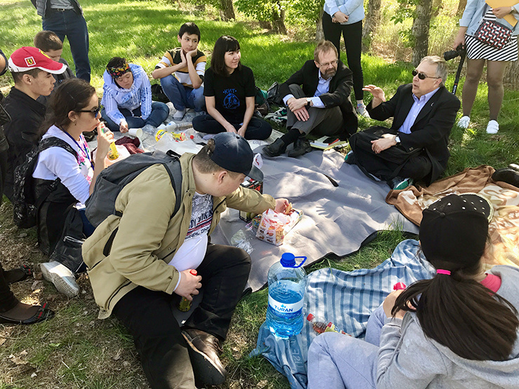 A group of people sit in a circle, having a picnic, while eating and listening to a man speak.