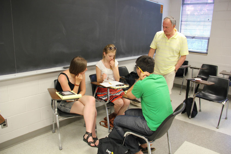 Instructor Alan Cherchesov talks to three Between the Lines participants, who are sitting in desks and writing.