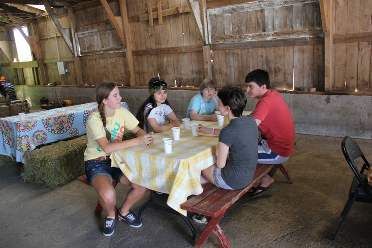 A group of Between the Lines participants sit at a picnic table in a barn at ZJ Farm in Solon, Iowa.