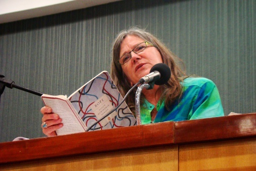 A woman sits at a panel and reads into a microphone from a book.