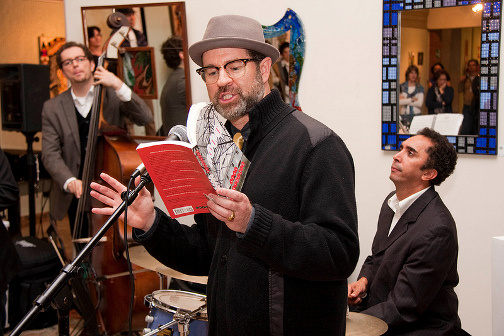 A person speaks into a microphone while people playing instruments can be seen in the background.