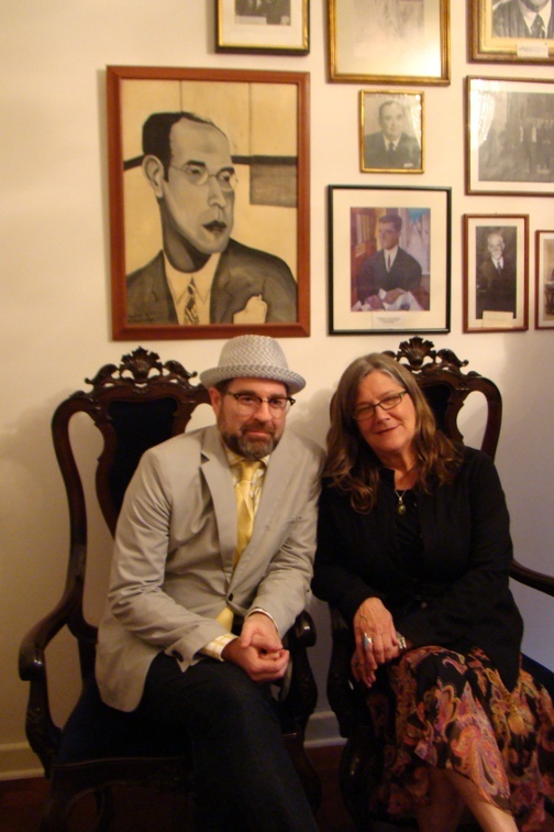 Two people sit in chairs and pose for a photo in front of a wall of painted portraits.