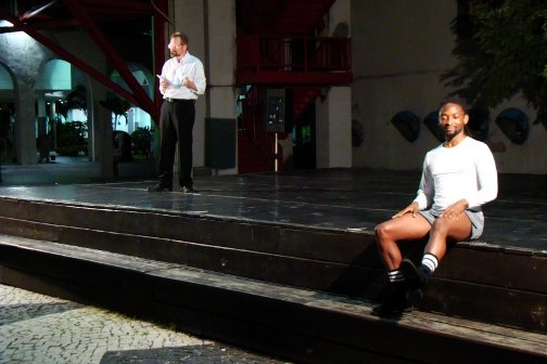 Chris Merrill reads from a paper on stage while another person sits to his left.