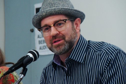 A person wearing a fedora and a striped button up shirt speaks into a microphone.