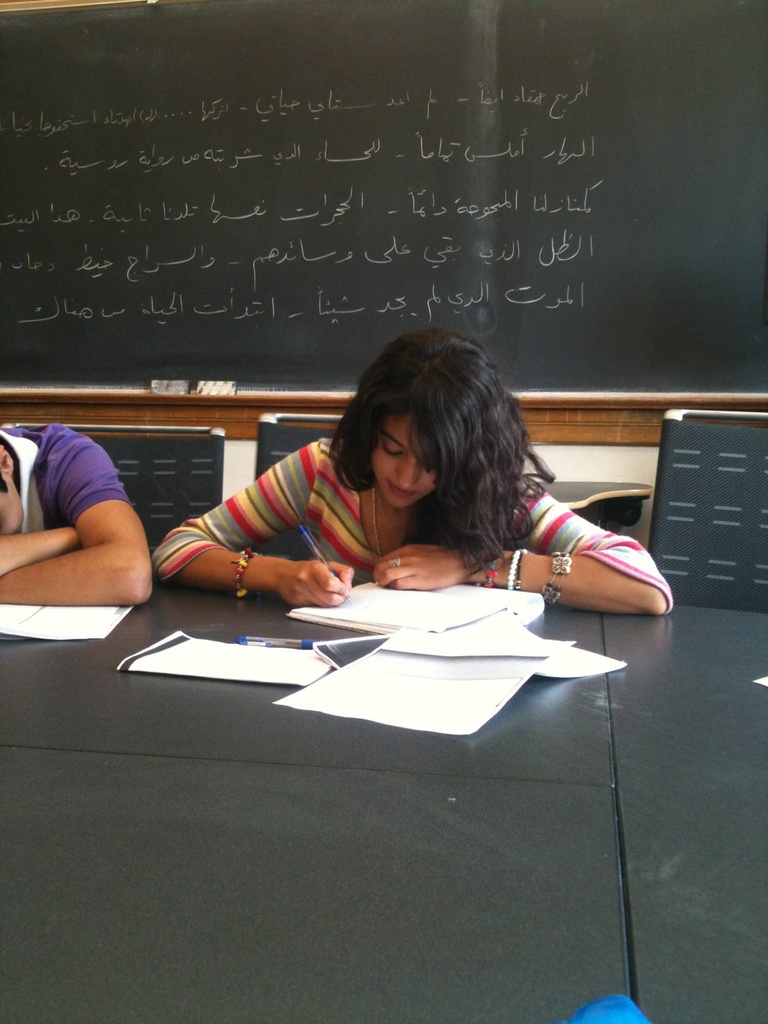 A Between the Lines participants writes in a notebook at a desk with a chalkboard with Arabic writing behind her.